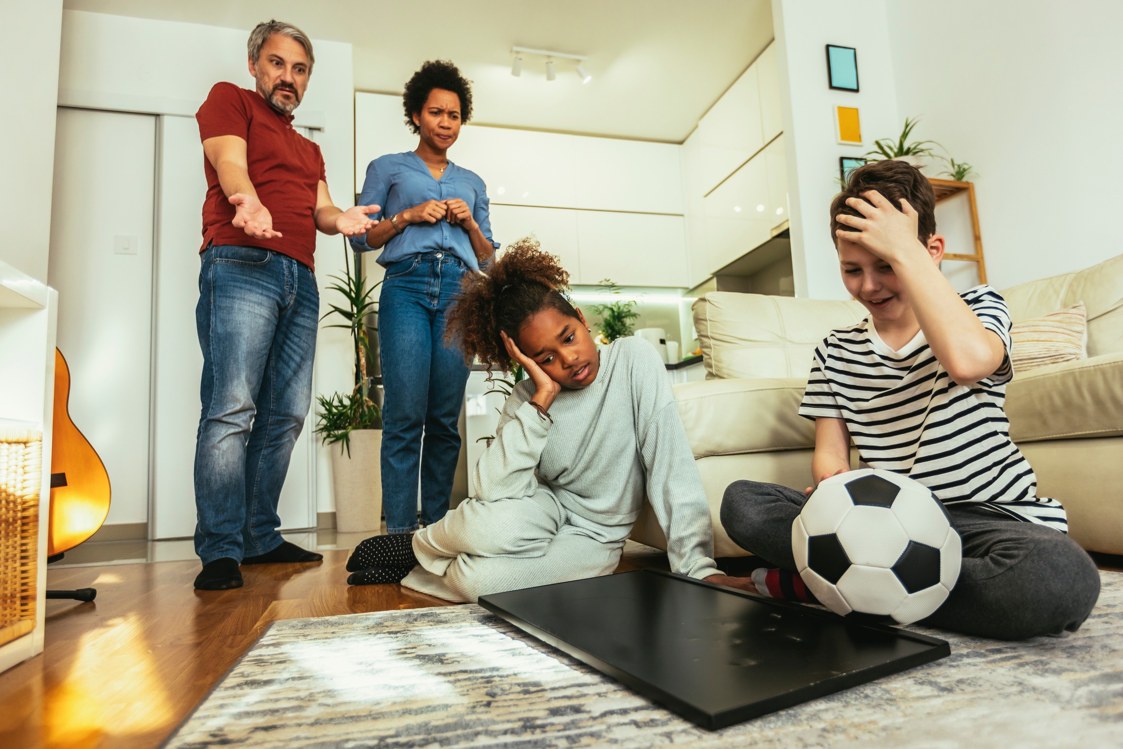 Das Bild zeigt zwei Kinder, die im Wohnzimmer vor einem kaputten Fernseher sitzen auf den ein Fußball gefallen ist, die Eltern stehen im Hintergrund.  