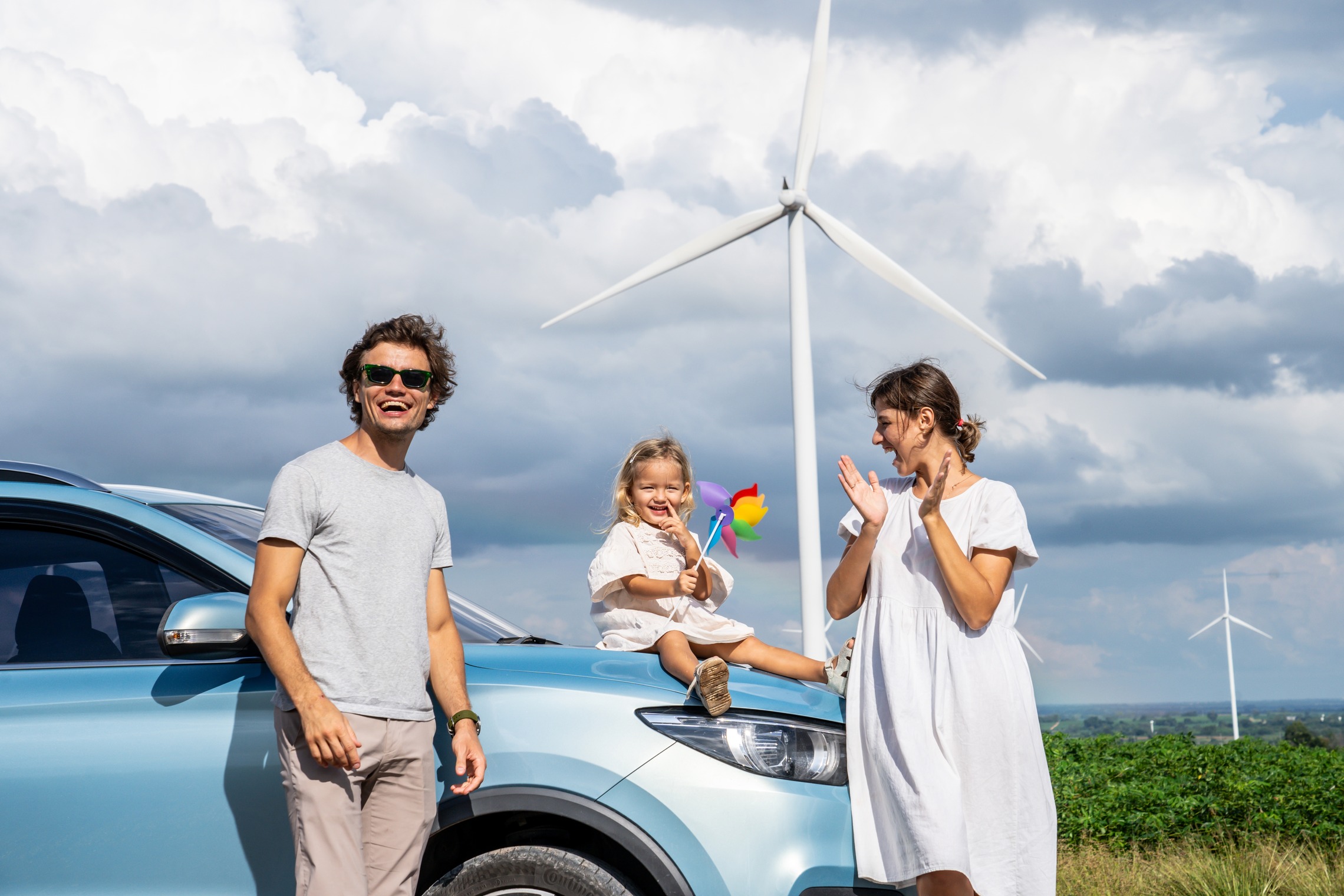 Das Bild zeigt eine Familie, die mit einem Auto vor einem Windrad steht. 