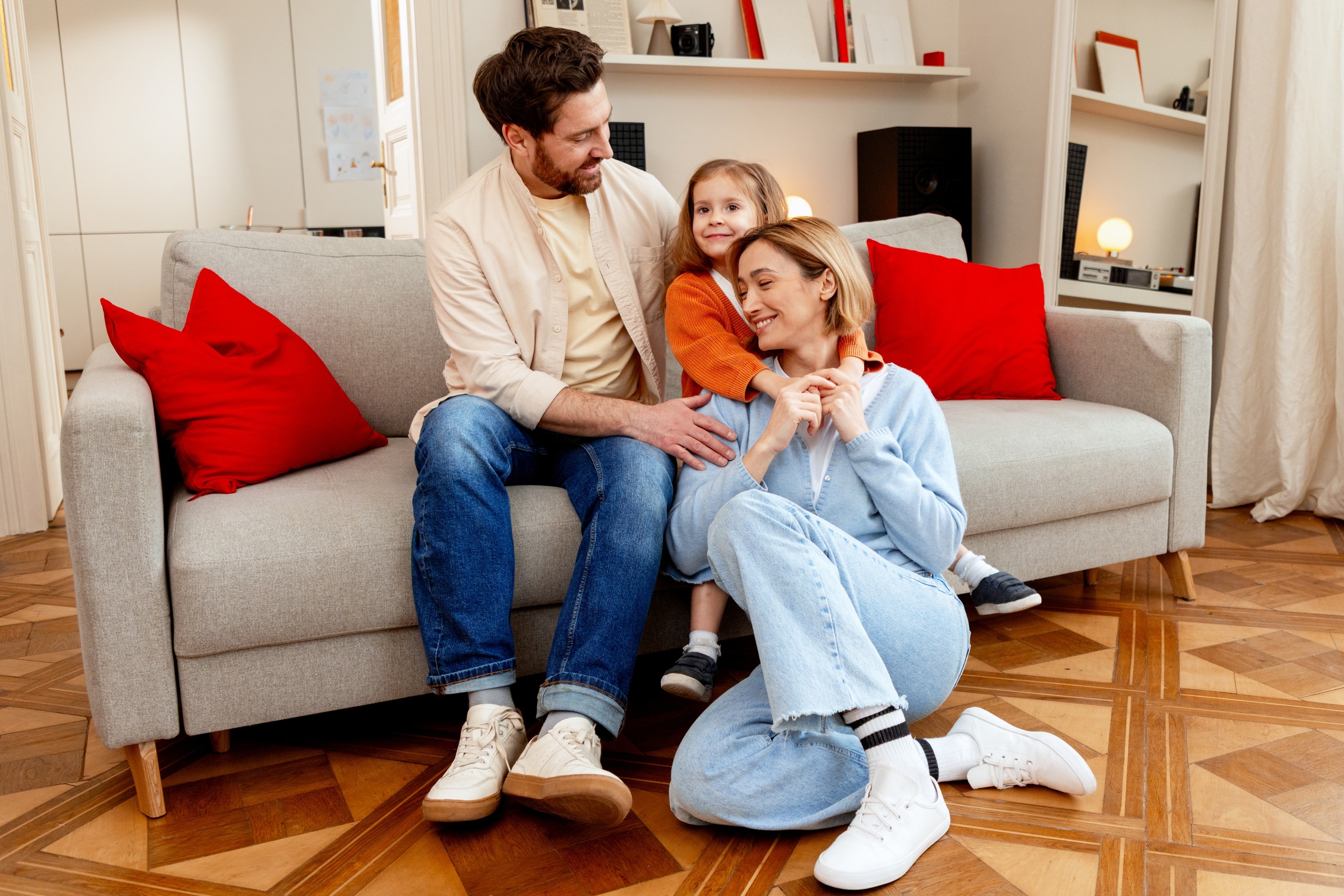 Das Bild zeigt einen Mann und ein Kind, das auf dem Sofa sitzt und eine Mutter, die darunter kniet.